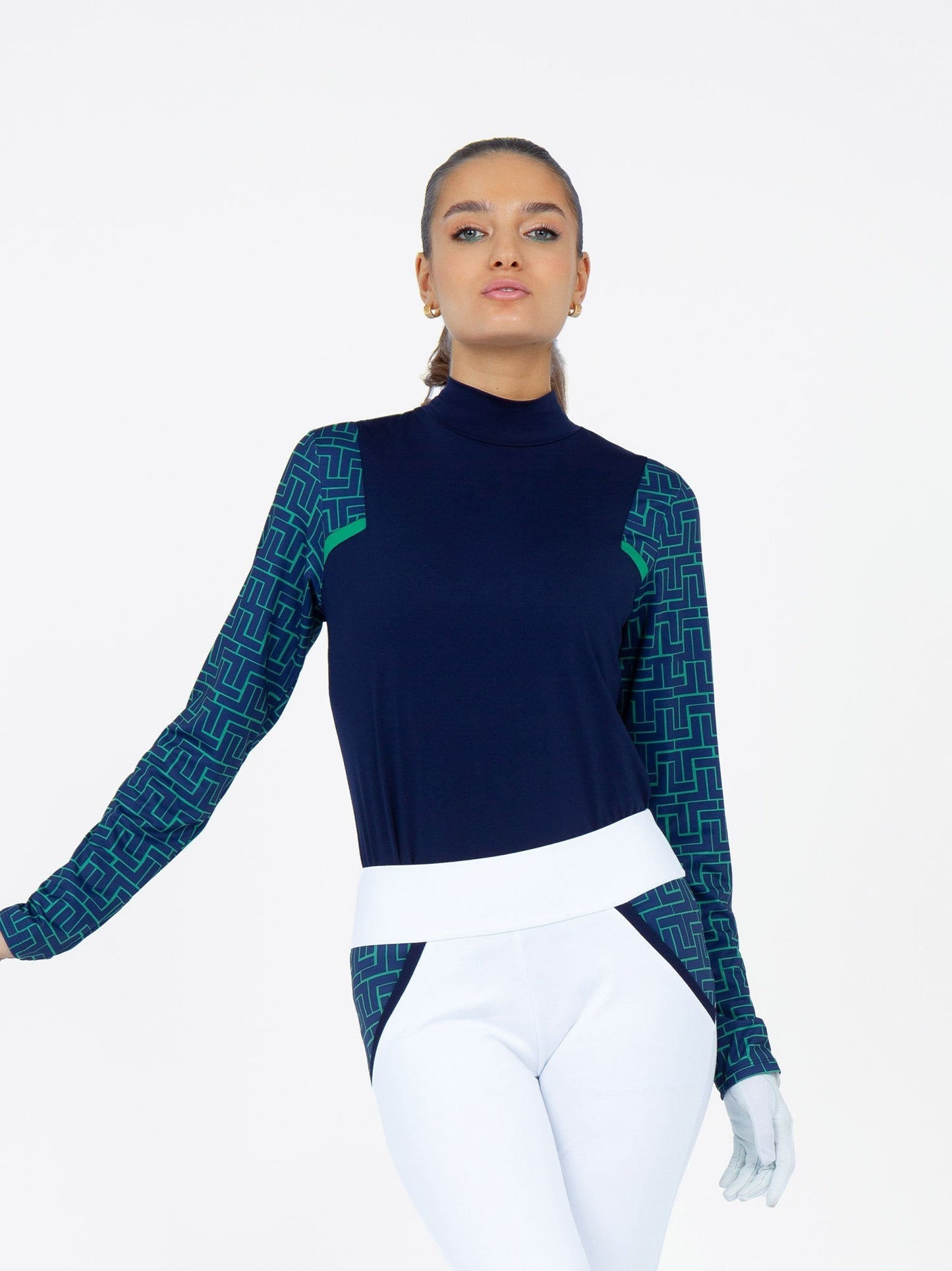 A woman stands against a white background wearing the inPhorm NYC Colorblock Active Sofia Long Sleeves Mock Neck in Ink/Ink Tessel with matching pants. Her hair is pulled back, she's confident, and wears a glove on her right hand.