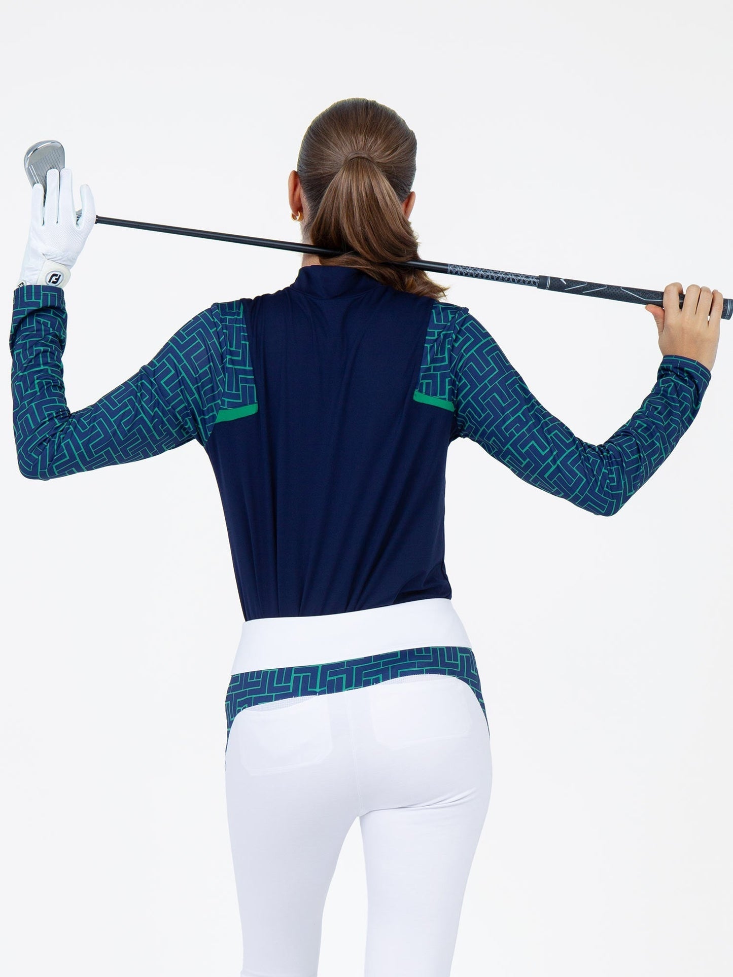 A woman with a ponytail faces away, holding a golf club behind her shoulders. She wears inPhorm NYC’s Colorblock Active Sofia Long Sleeves Mock Neck in Ink/Ink Tessel, white gloves, and fitted white pants against a plain white background.