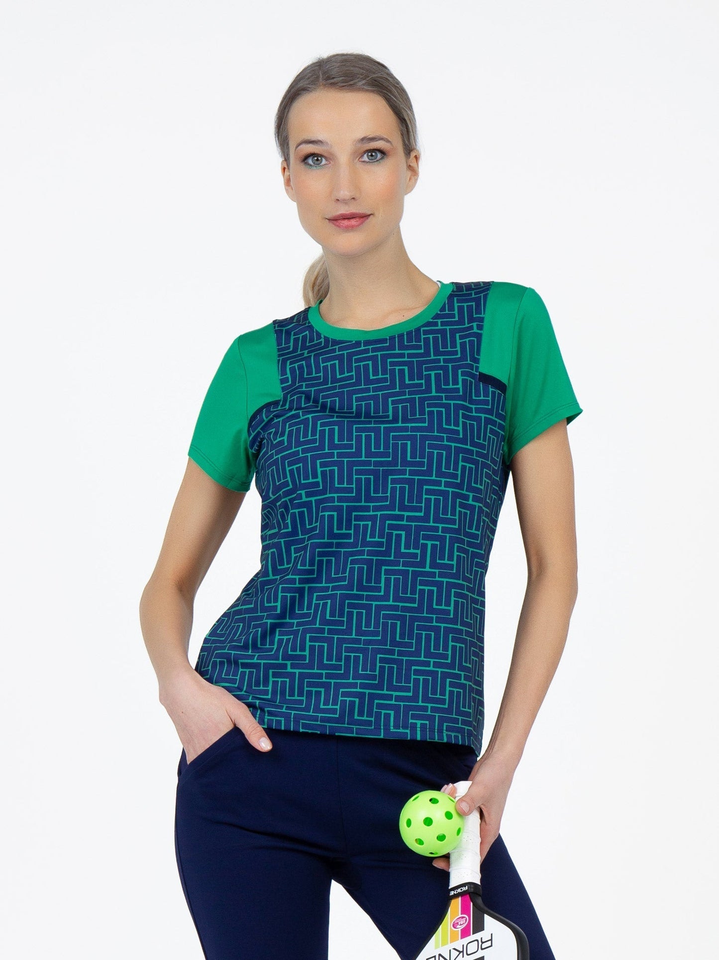 A woman with light skin and blue eyes poses against a white background, wearing the inPhorm NYC Colorblock Active Sofia Short Sleeve Crew Neck in Ink Tessel/Ivy. She holds a white and pink paddle and a green perforated ball, gazing confidently at the camera.