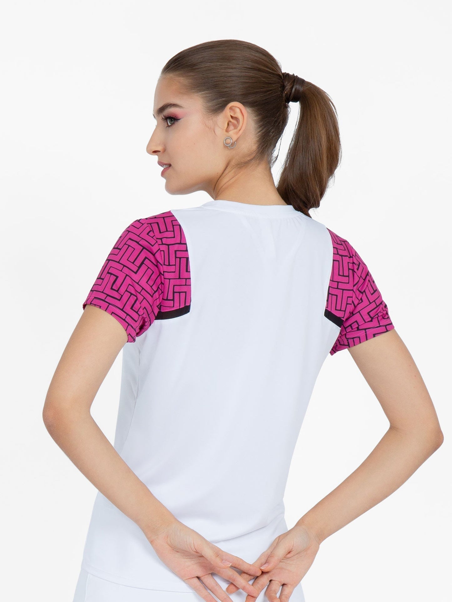 A woman with light skin and brown hair in a ponytail faces away from the camera, fingers clasped behind her, wearing the inPhorm NYC Colorblock Active Sofia Short Sleeve Crew Neck in Raspberry Pink. The background is plain white.