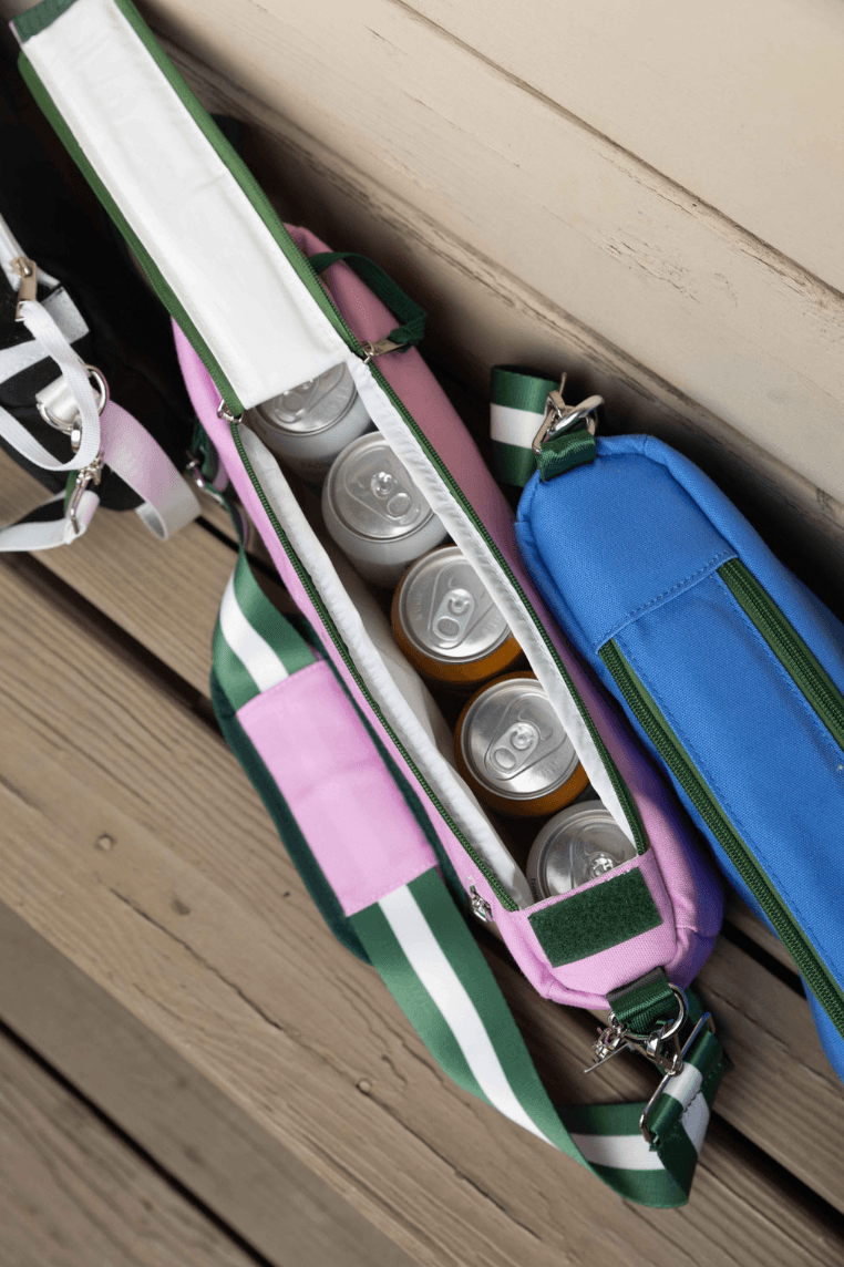 Three Fresh Pickle Insulated Cooler Bags with striped straps sit on wooden planks. The open pink bag shows several silver cans, while blue and green bags are nearby—perfect for keeping your essentials cool courtside.