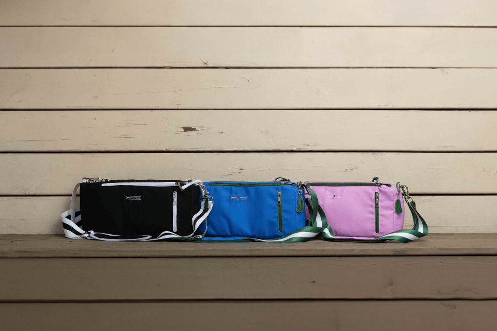 Three Fresh Pickle Insulated Cooler Bags in black, blue, and pink sit side by side on a wooden bench. Each soft-sided bag has a zip closure, front label, and adjustable strap—perfect for court necessities in style.