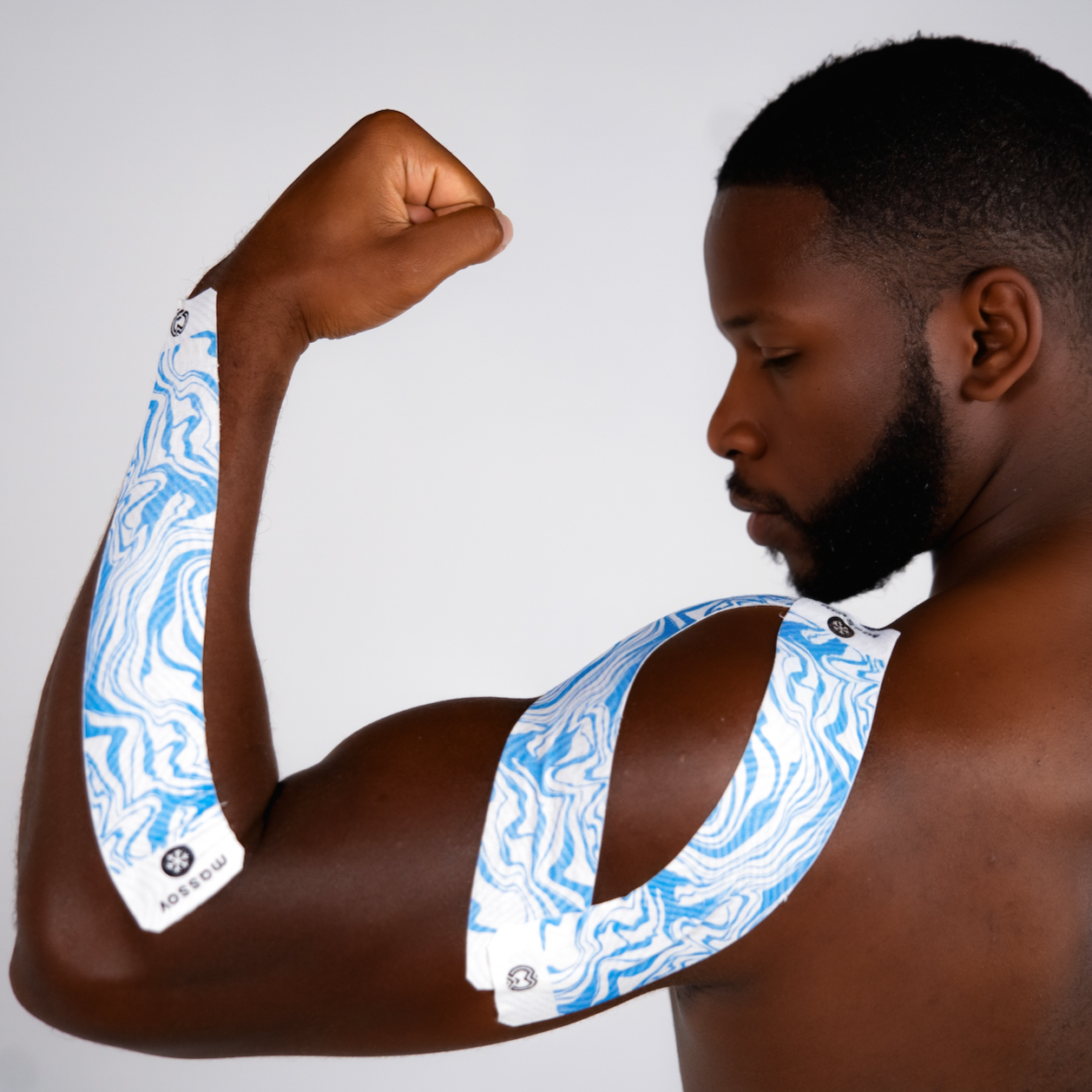 A muscular man flexes his right arm, showing Massov Cooling Menthol Kinesiology Tape in a blue and white pattern applied from shoulder to forearm. The tape supports muscles and helps prevent injury. The background is plain and light.