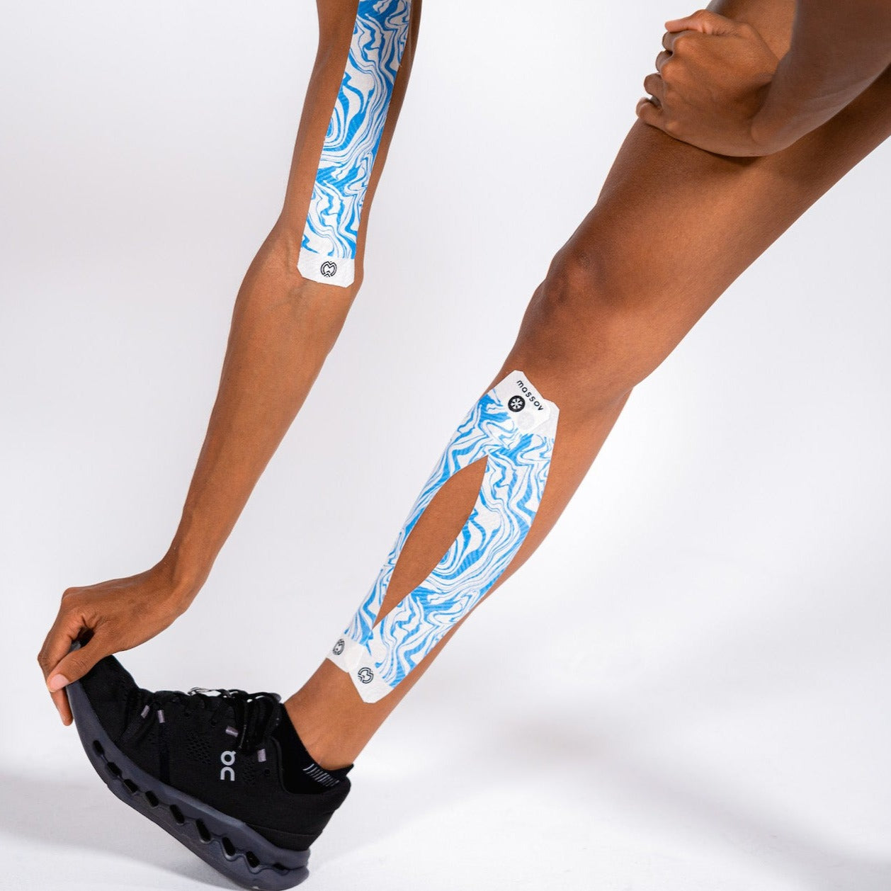 Wearing black athletic shoes, a person bends forward with Massov Cooling Menthol Kinesiology Tape applied to their forearm and lower leg. The blue-and-white tape stands out against the plain white background, highlighting the athletic pose.