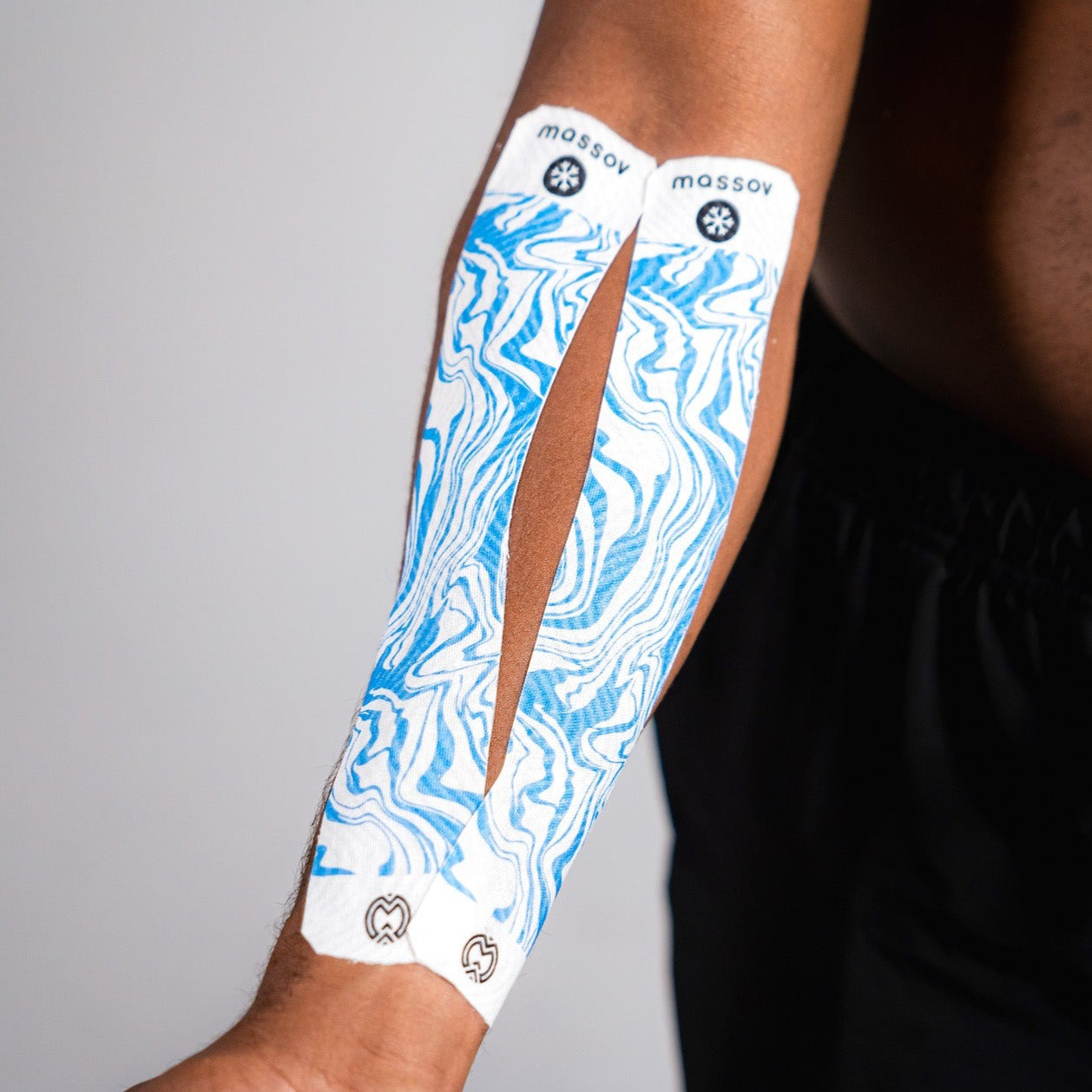 A forearm with two white and blue marbled Massov Cooling Menthol Kinesiology Tapes applied from below the elbow to the wrist, supporting muscle recovery. The background is plain, and the person is wearing black shorts.