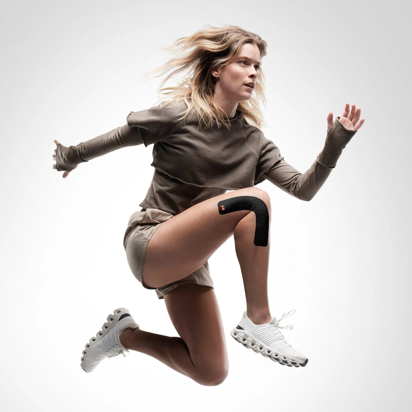 A blonde woman in a taupe long-sleeve shirt, matching shorts, and white athletic shoes jumps against a white background, KT Tape Starter Pack by KT Tape supporting her right knee with black kinesiology tape for pain relief.