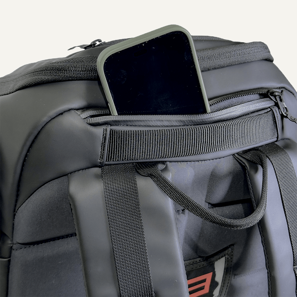 A black smartphone peeks out of a small zippered pocket on the FORWRD Court Caddy Pickleball Bag, which features padded straps, a sturdy handle, and smooth matte fabric with visible stitching against a plain background.