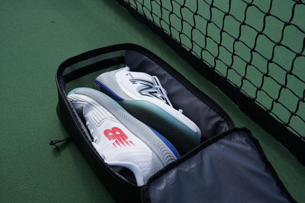 A pair of white sneakers with red and blue accents are neatly stored inside the black FORWRD Court Caddy Shoe Cube, resting on a green tennis court beside a black net.