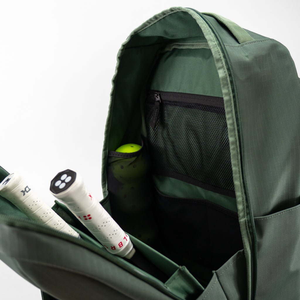 A close-up of the FORWRD Court Ranger Pickleball Backpack shows two racquet handles, a pickleball, and a mesh zip pocket inside. The durable green bag is featured against a plain white background.