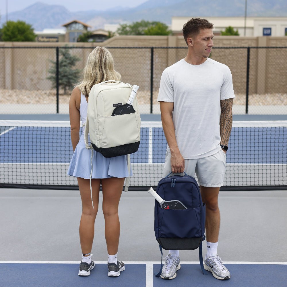 A woman faces away on a tennis court, wearing athletic gear and holding the FORWRD Court Ranger Pickleball Backpack, while a man beside her holds a dark pickleball bag with rackets. Both are in sneakers; a net and buildings appear in the background.