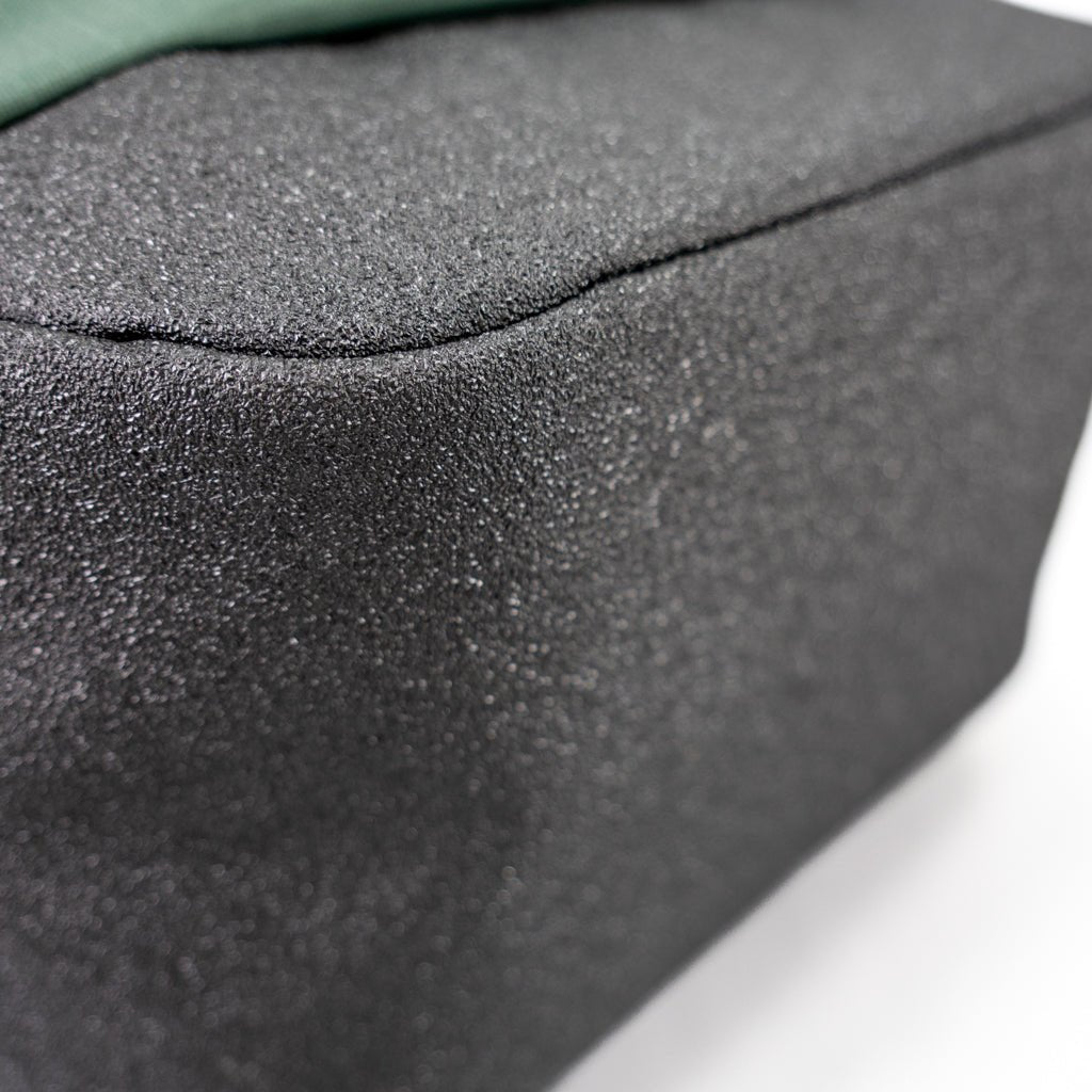 Close-up of a textured black FORWRD Court Ranger Pickleball Backpack with slightly rounded edges on a white surface. The material looks rough and grainy, with a hint of green fabric at the top edge.