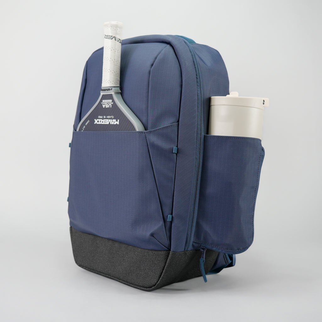 The FORWRD Court Ranger Pickleball Backpack stands upright with a white paddle in the front pocket and a large tumbler in the side pocket, making it one of the best pickleball bags under $200. The background is plain and light-colored.