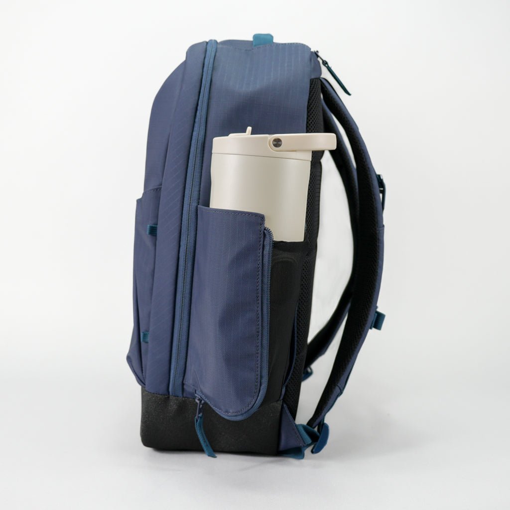 The FORWRD Court Ranger Pickleball Backpack in navy blue is shown side view against a white background, featuring a beige travel mug in its side pocket. The bag includes multiple zippers, padded back, and adjustable shoulder straps.