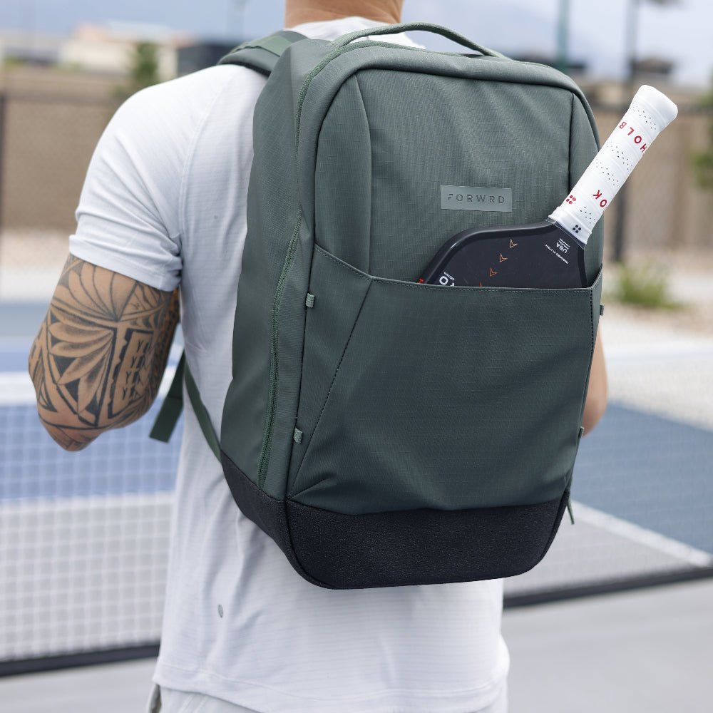 A person with a tattooed arm wears a light gray shirt and carries the FORWRD Court Ranger Pickleball Backpack, with a black paddle and white tube of balls visible from the front pocket, standing on an outdoor pickleball court.