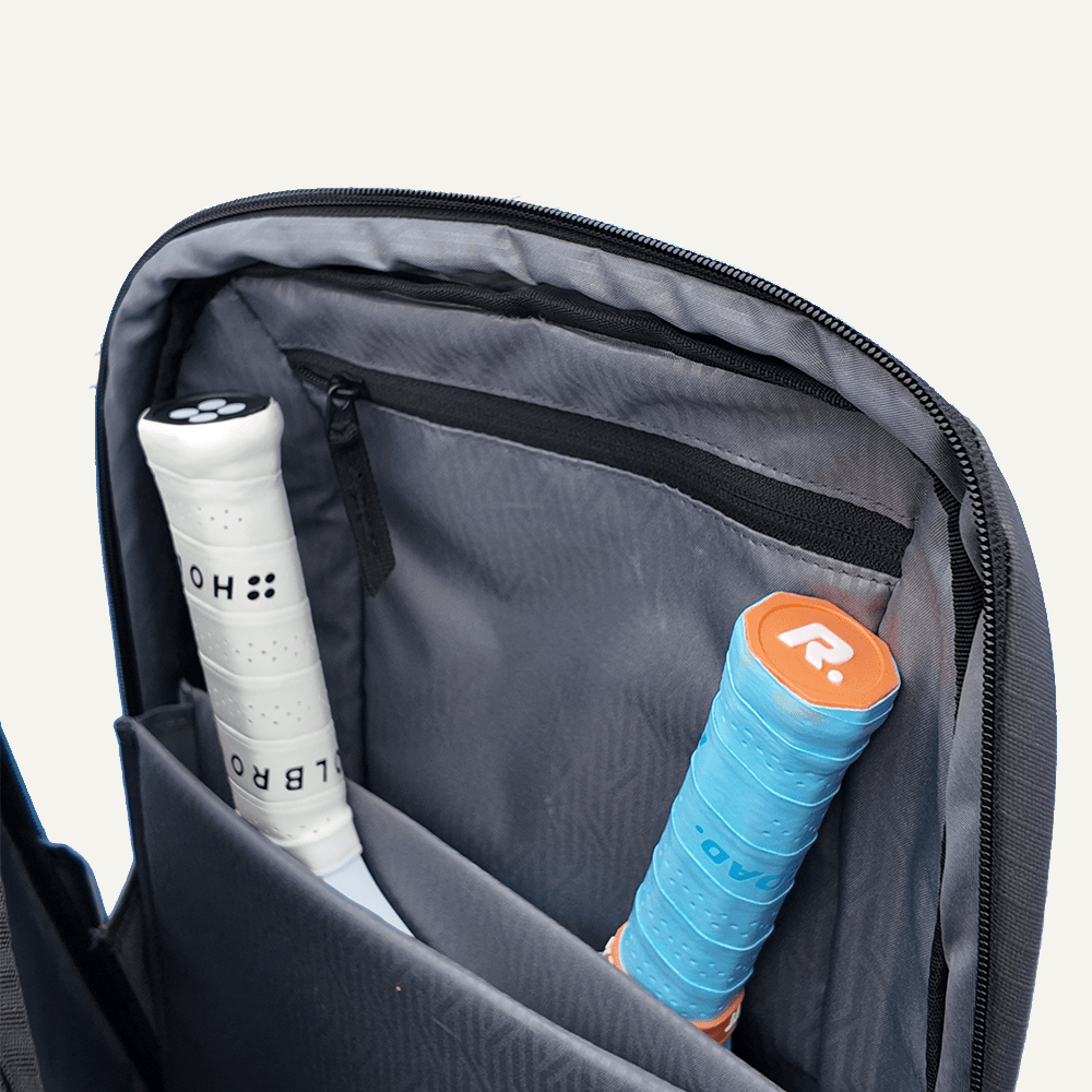 The FORWRD Court Ranger Pickleball Backpack's gray interior, with a padded divider like those in premium bags, is shown open with two rackets inside—one white-gripped with black accents and another with a blue grip and orange end cap.