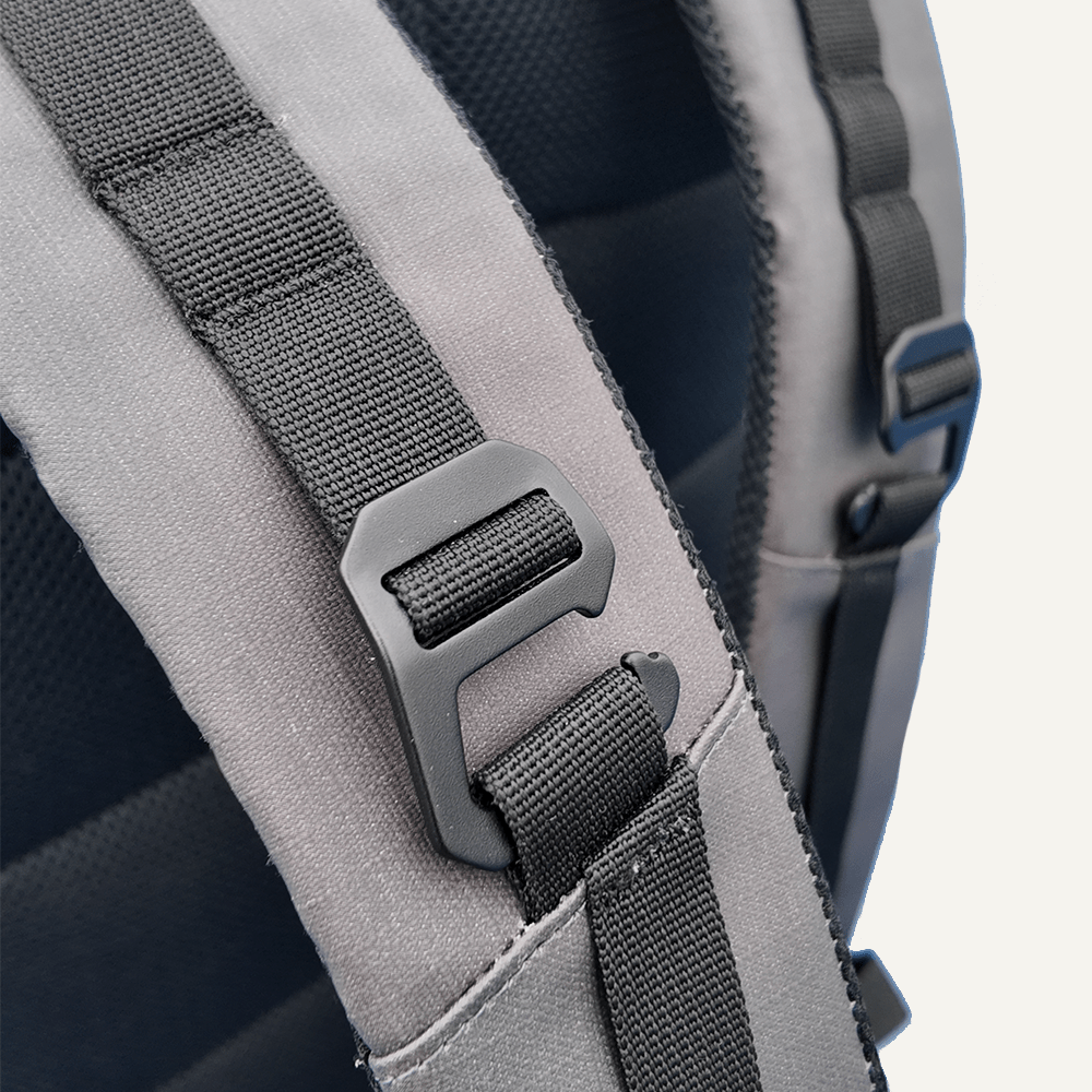 Close-up of a black plastic buckle on a black nylon strap, attached to the grey padded shoulder strap of the FORWRD Court Ranger Pickleball Backpack. Visible stitching and a blurred, light-colored background complete the scene.