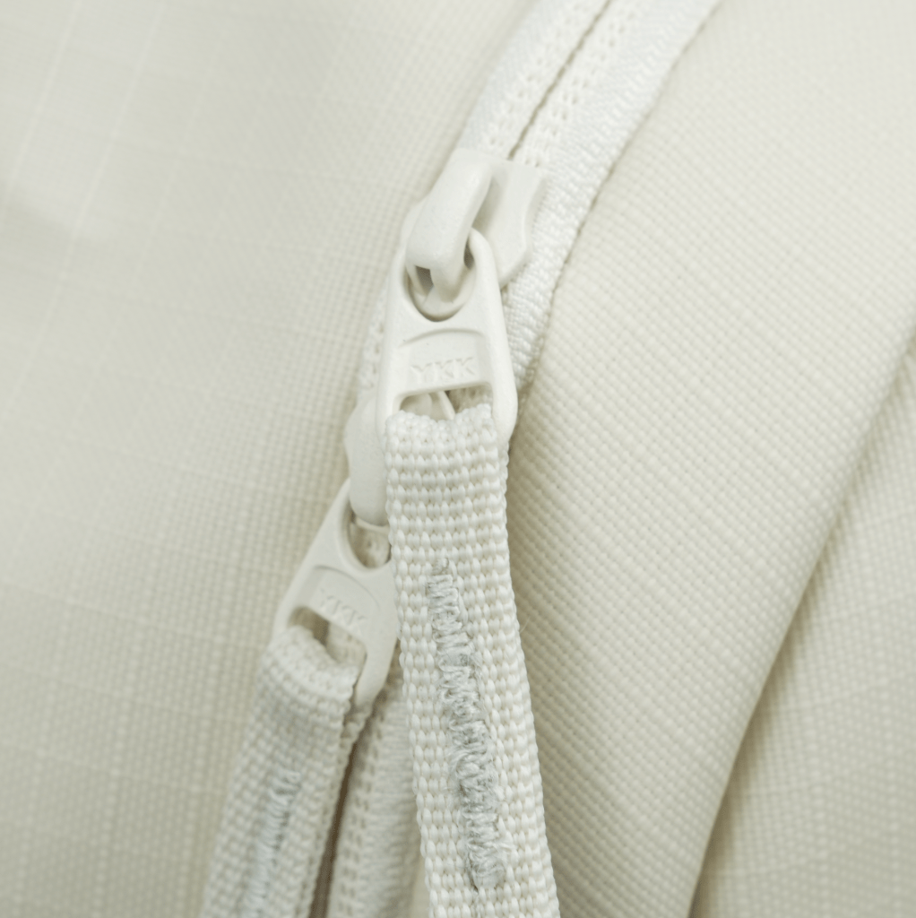 Close-up of two white zippers with knotted fabric pulls on the FORWRD Court Ranger Pickleball Backpack. The light, textured fabric features a subtle grid pattern, and parallel zippers show visible stitching.