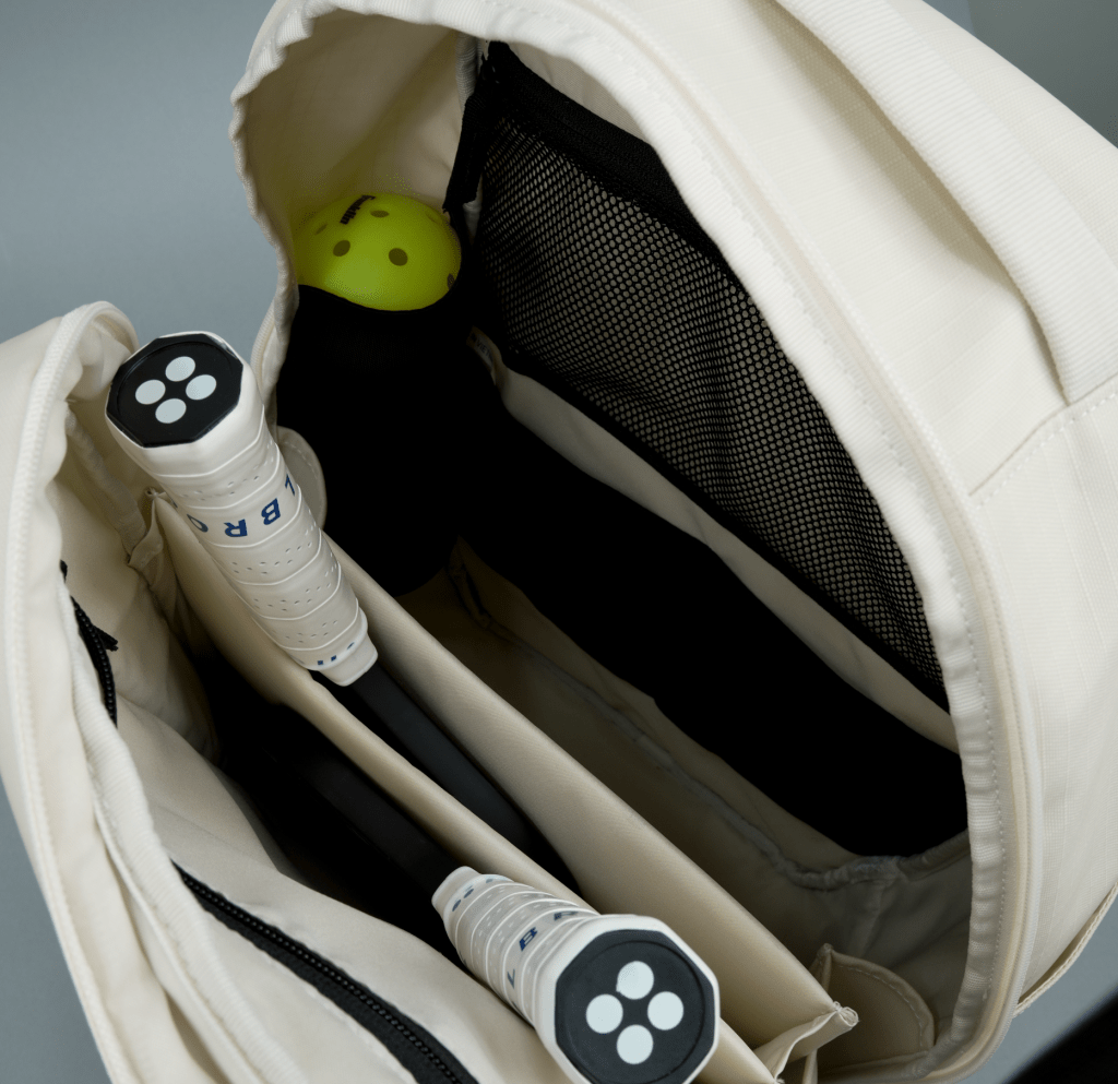 The FORWRD Court Ranger Pickleball Backpack in cream is unzipped, showing two paddles with white handles and a yellow perforated ball inside. The paddles are stored in a padded section next to a black mesh interior pocket.