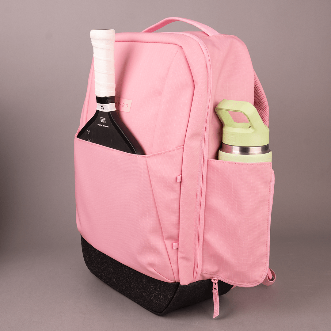 The FORWRD Court Ranger Pickleball Backpack in pink features a front pocket for your paddle, side pocket for a water bottle, and sits against a gray background—a stylish, on-the-go essential for pickleball players.