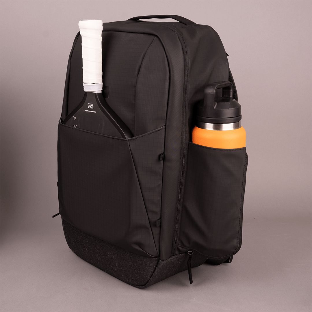 The FORWRD Court Ranger Pickleball Backpack stands upright on a neutral background, with a tennis racket handle in the back pocket and an orange water bottle in the side pocket. Its sleek, structured design features multiple compartments.