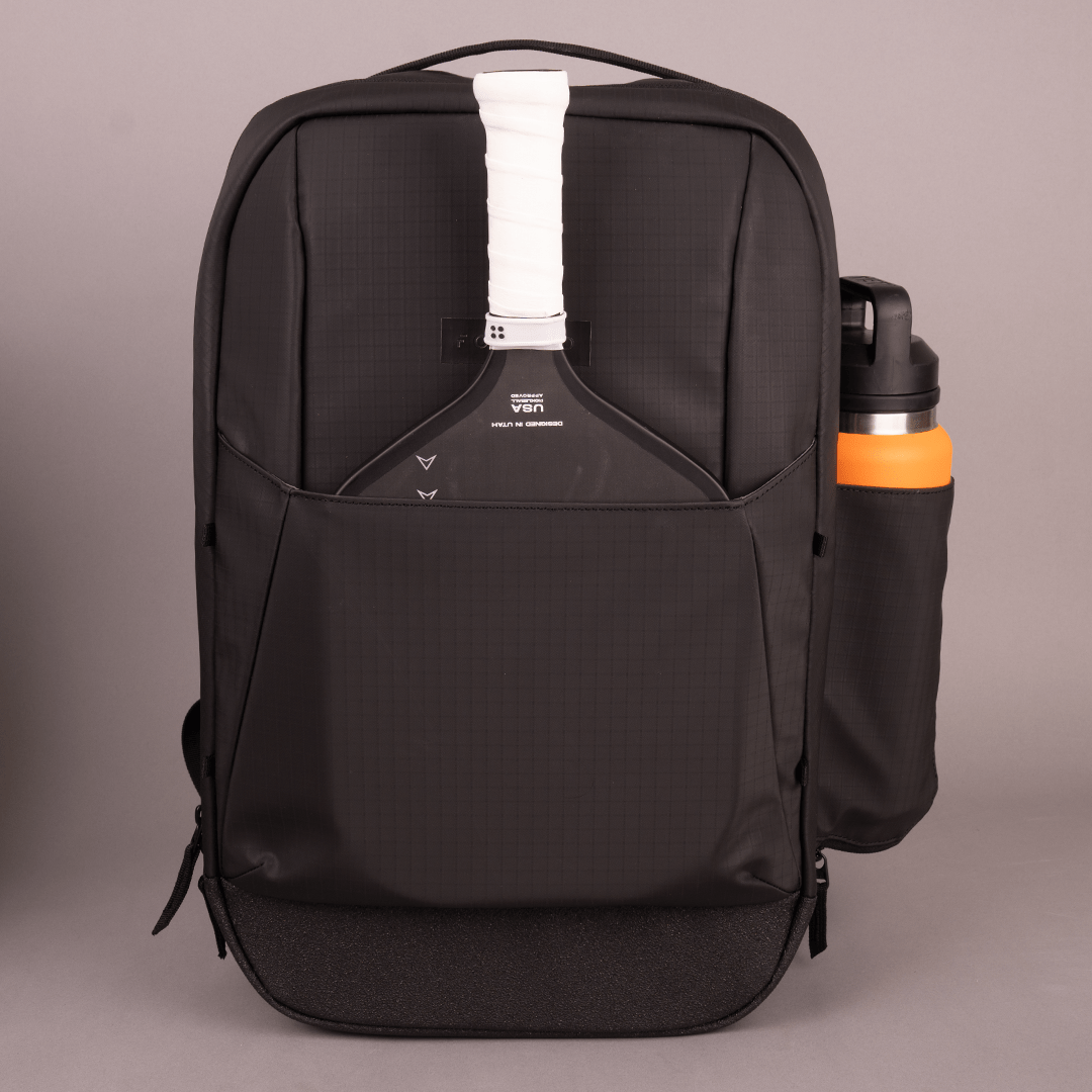 The FORWRD Court Ranger Pickleball Backpack stands upright and forward-facing, with a white-handled tennis racket in the front pocket and a black-and-orange water bottle in the side holder, set against a plain gray background.