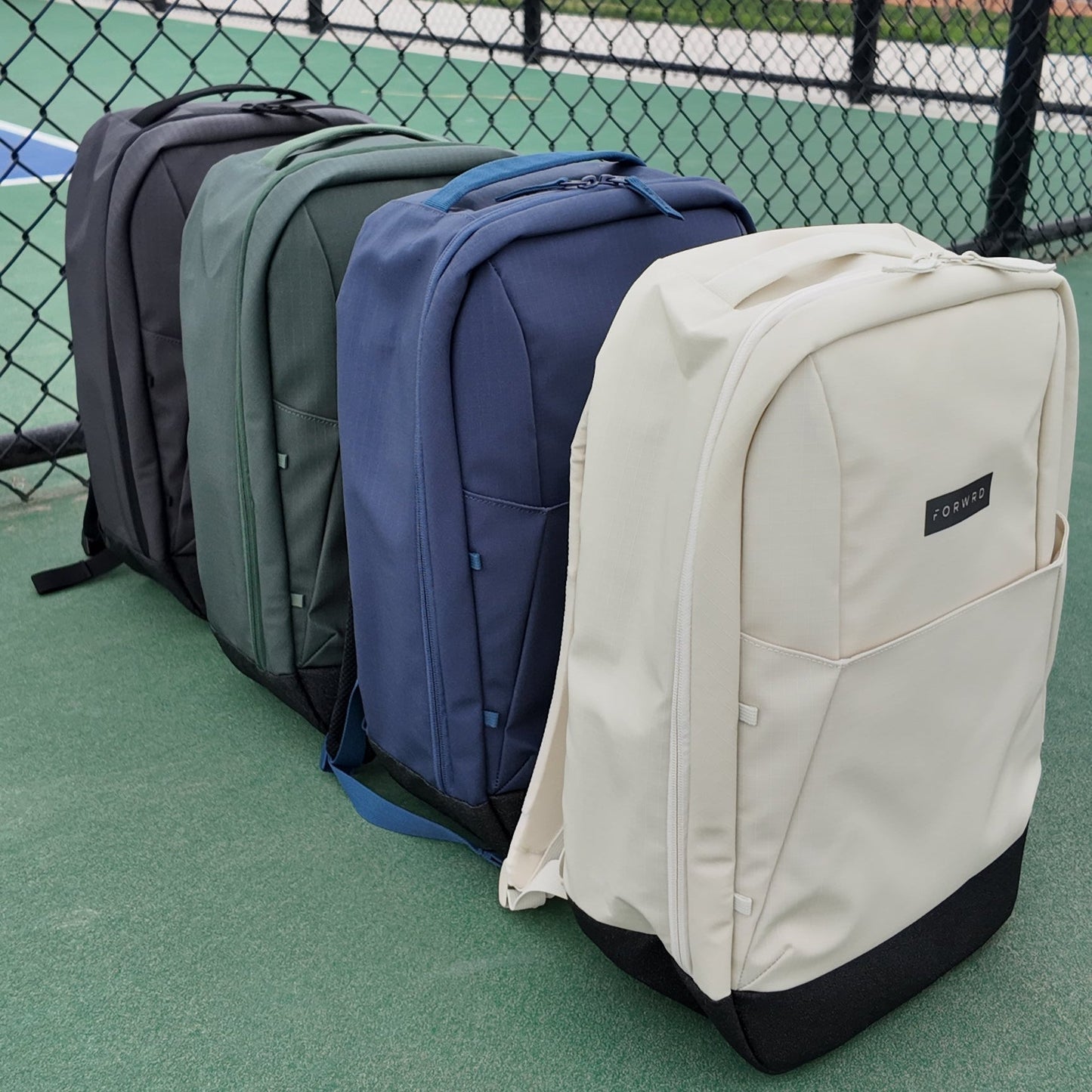 Four FORWRD Court Ranger Pickleball Backpacks in black, dark green, navy blue, and off-white are lined up on a green tennis court. The off-white style offers a chic pickleball bag option for players on the move.