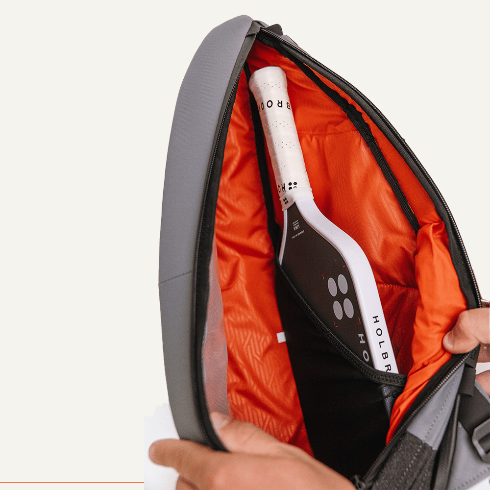 A close-up of the FORWRD Cross Court Pickleball Sling shows its black exterior and bright orange lining. Inside, a hand reveals a white-handled HOLBR paddle neatly stored in an inner pocket—ideal for lightweight, durable storage on the go.
