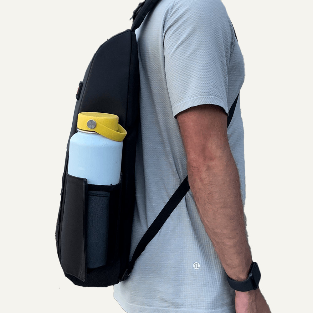 A person with a light gray short-sleeve shirt and black smartwatch carries the FORWRD Cross Court Pickleball Sling. A large white water bottle with a yellow handle is in the side pocket. The background is plain and light-colored.