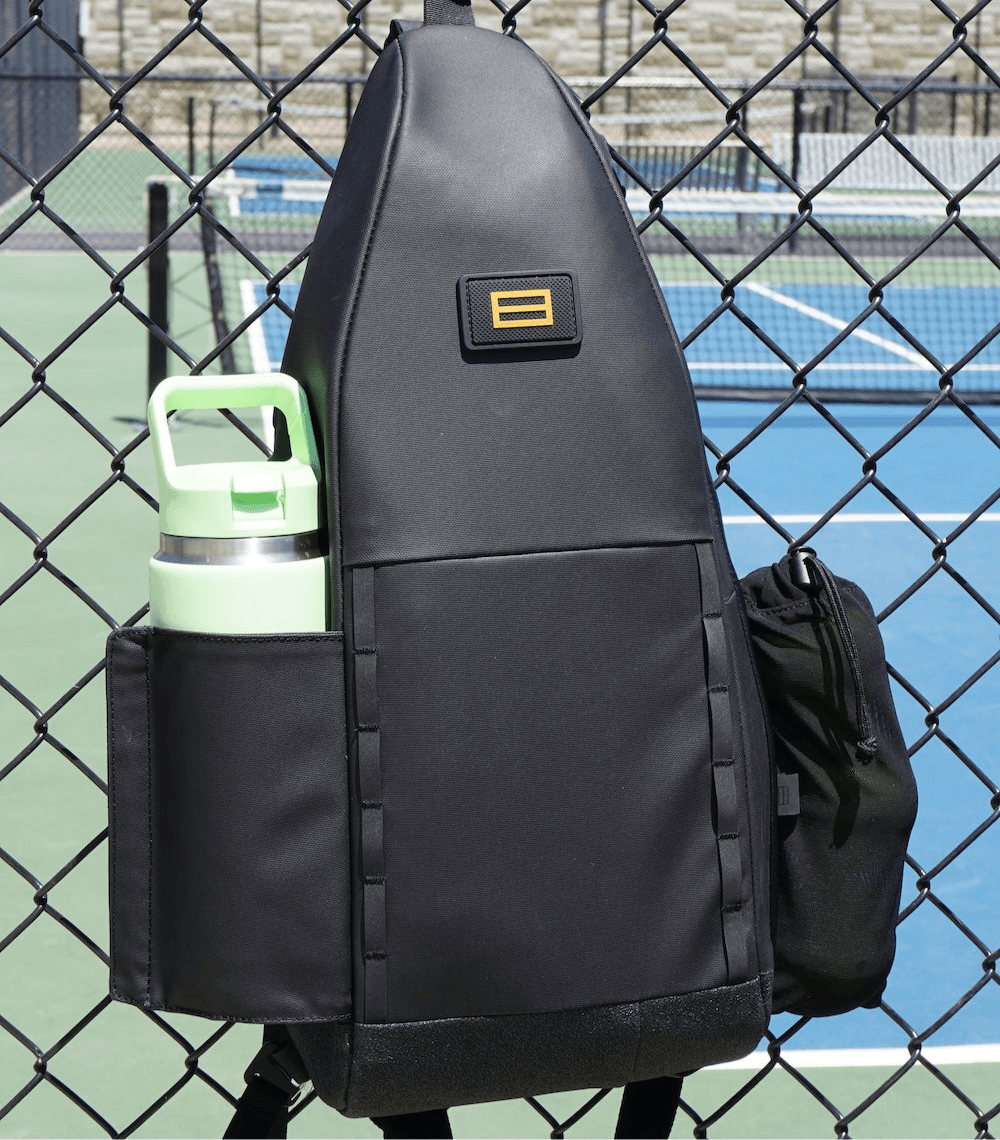 The FORWRD Cross Court Pickleball Sling, in sleek black with a yellow logo, hangs on a chain-link fence by the court. It features a side pocket for a mint green water bottle, another with a black pouch, and is lightweight yet durable on any blue-green court.