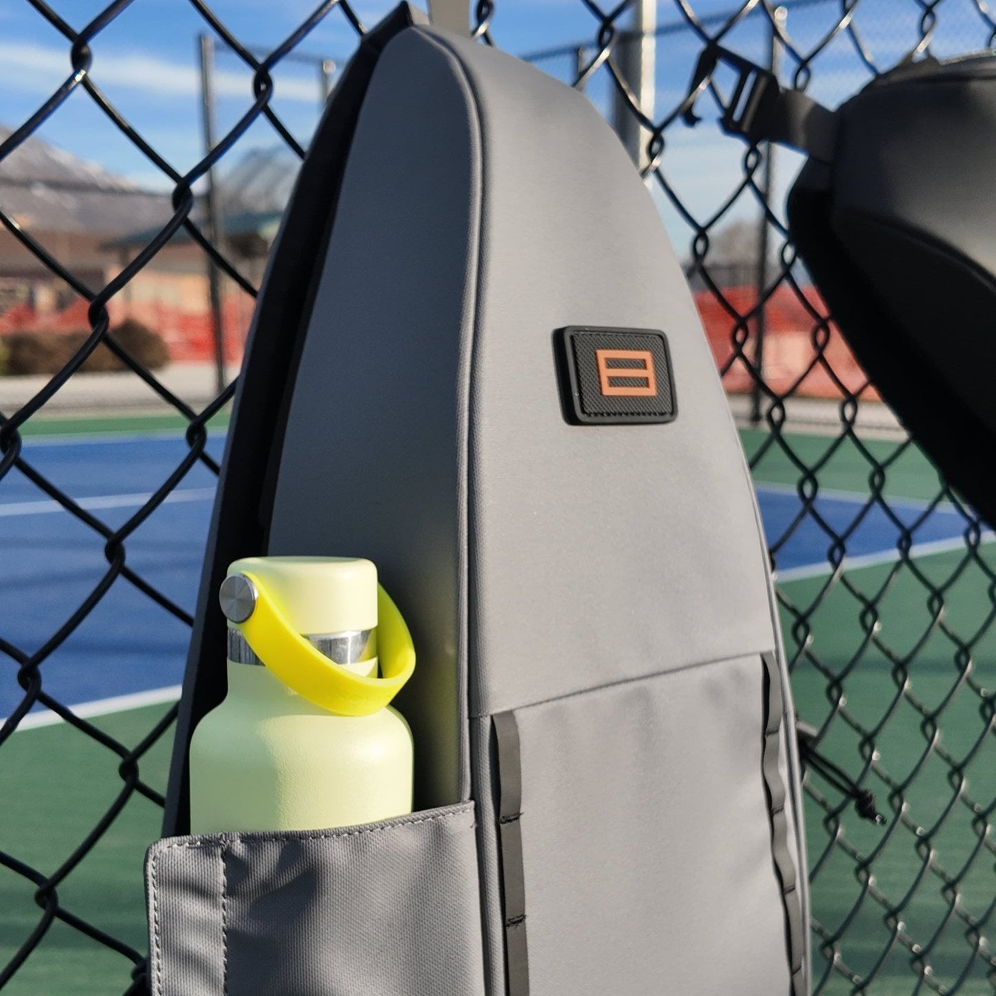 The FORWRD Cross Court Pickleball Sling hangs on a chain-link fence at an outdoor court, holding a light yellow water bottle with a yellow handle in its side pocket. Blurred mountains and trees appear in the sunny background.