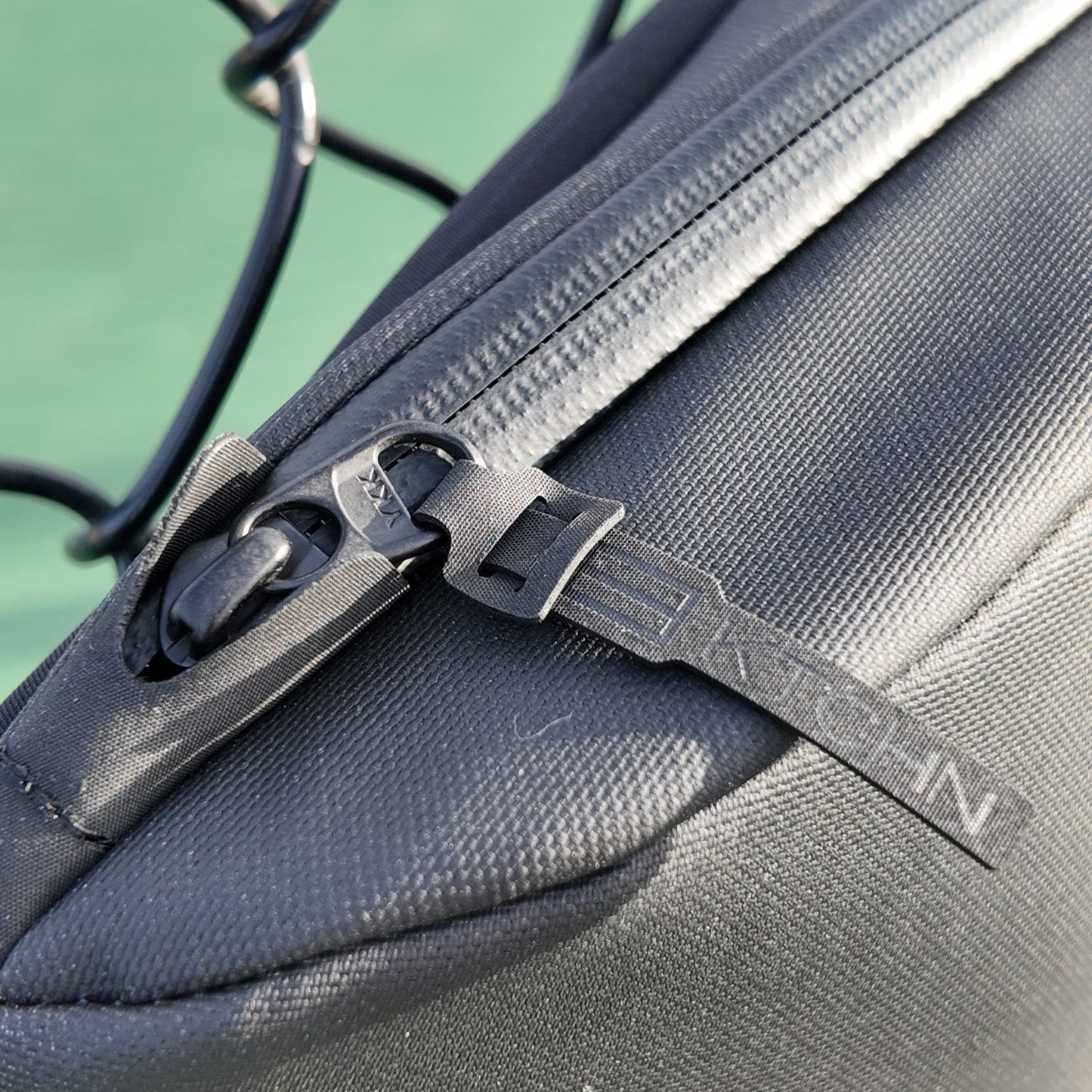 Close-up of a black FORWRD Cross Court Pickleball Sling with visible zipper and branded pull. The blurred outdoor background with a wire fence and green surface suits this lightweight, durable pickleball bag.