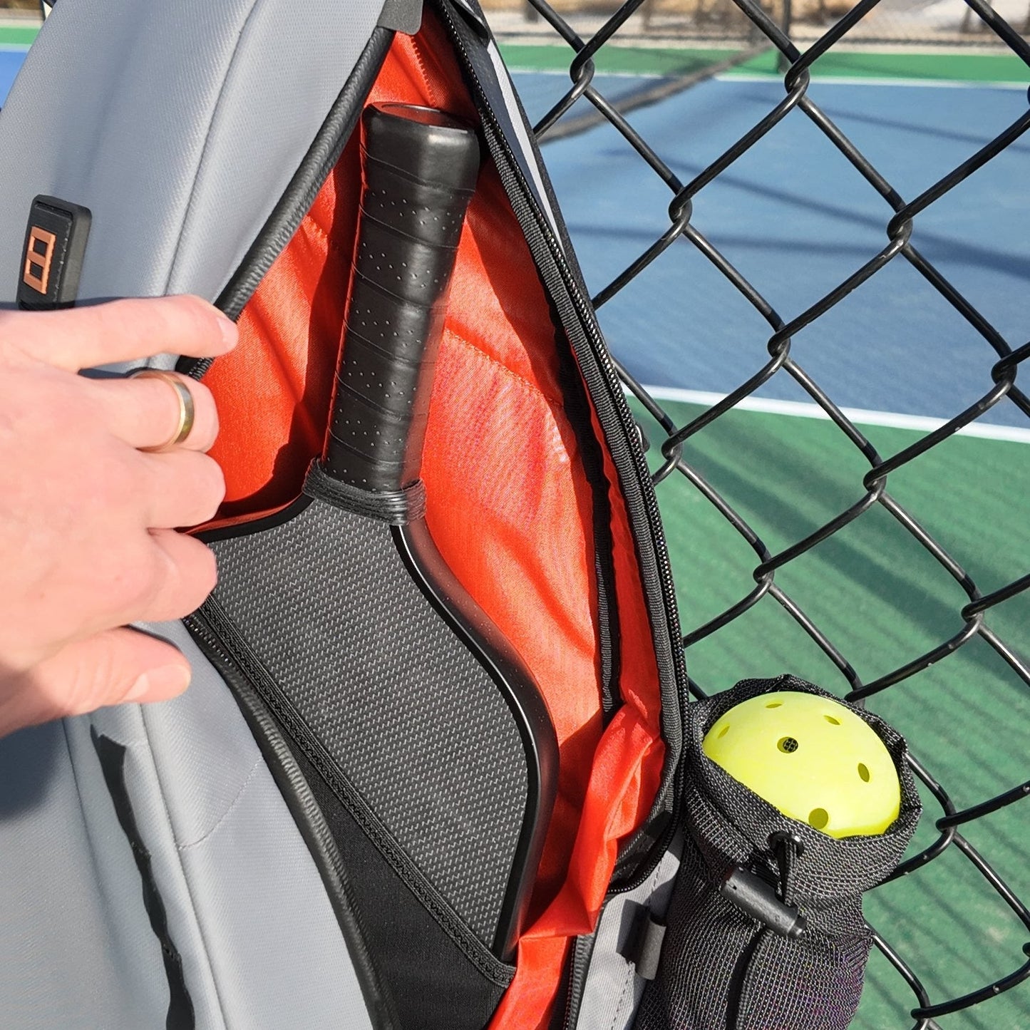 A hand opens the FORWRD Cross Court Pickleball Sling, revealing a black paddle inside. The lightweight, durable gray and red bag hangs on a fence by a court, with a yellow pickleball in its mesh holder as sunlight casts shadows.