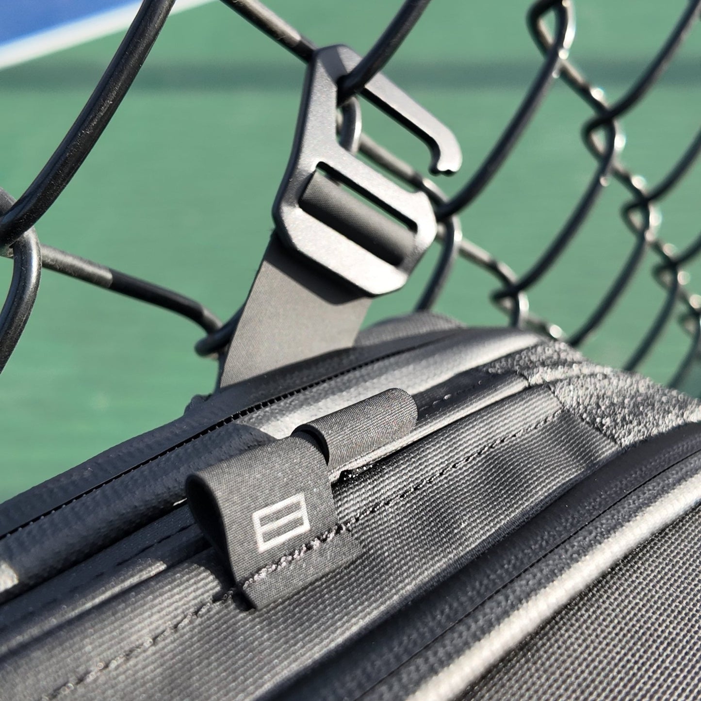Close-up of the FORWRD Cross Court Pickleball Sling, a lightweight, durable bag with a zipper and rectangular tag, hanging from a matte black carabiner on a fence. Sunlit green pickleball court and blue line appear blurred in the background.