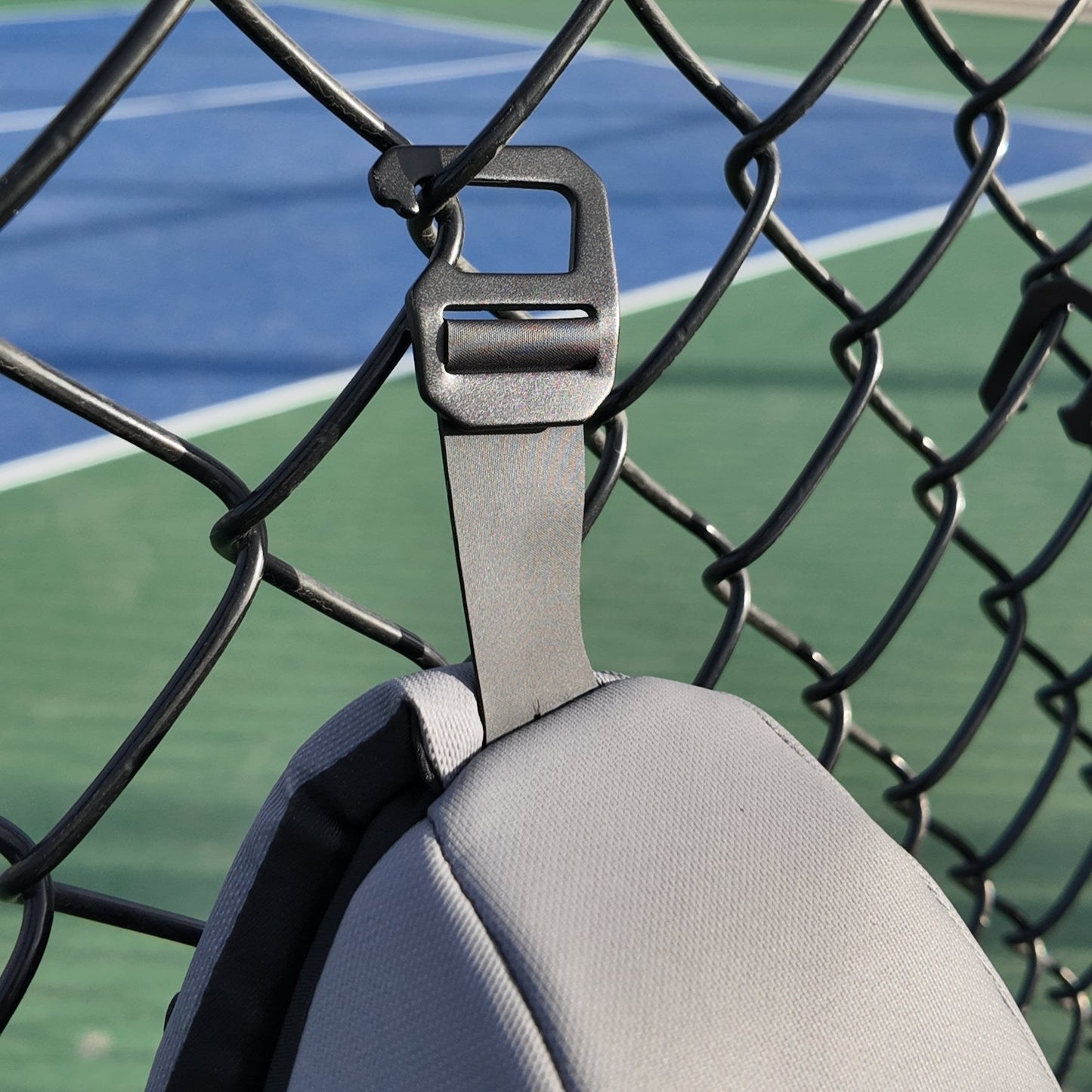 The FORWRD Cross Court Pickleball Sling hangs on a black hook attached to a chain-link fence, with sunlight illuminating the blue and green tennis court in the background, creating an inviting outdoor sports vibe.