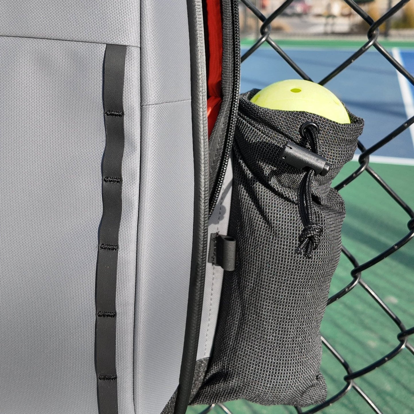 A close-up of the FORWRD Cross Court Pickleball Sling, a lightweight and durable bag, hanging on a chain-link fence. Its mesh side pocket holds a yellow ball, with a softly blurred blue and green court in the background.