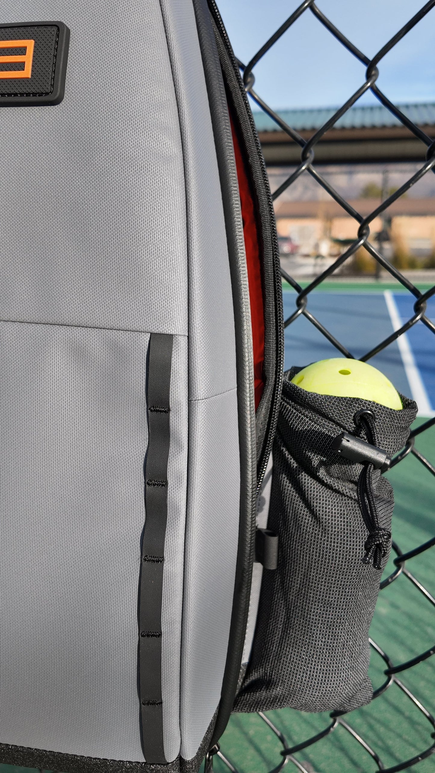A close-up of the FORWRD Cross Court Pickleball Sling, a lightweight and durable bag, hangs on a chain-link fence. Its outer mesh pocket holds a yellow pickleball. A blue court and building are blurred in the sunny background.