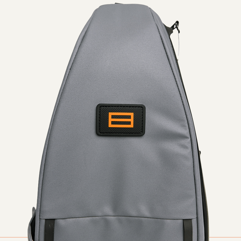 A close-up of the FORWRD Cross Court Pickleball Sling shows a black patch with an orange equal sign on the gray, smooth matte fabric. The bag features lightweight durability, and its zipper is partially visible on the right edge.
