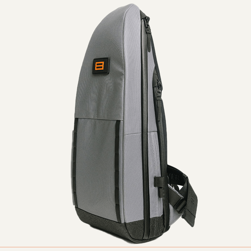 The FORWRD Cross Court Pickleball Sling is a modern gray backpack with black trim, orange rectangular logo, front pocket, side zipper, adjustable black textured strap, and an angular shape. Lightweight and durable, it stands upright.