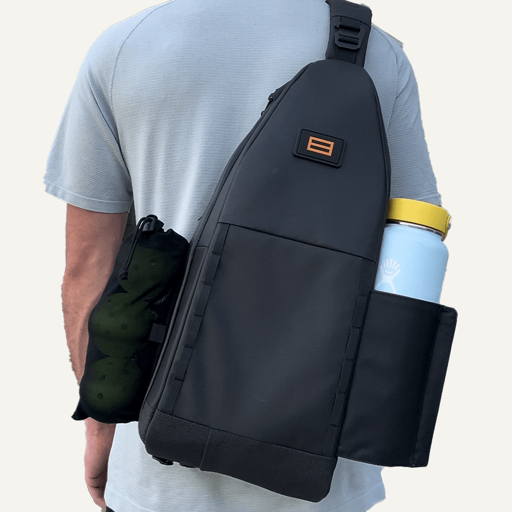 A person in a light grey shirt carries the FORWRD Cross Court Pickleball Sling—sleek, black, and lightweight—with a side pocket for a yellow-lid water bottle, a cinched strap pouch, and an orange logo patch.