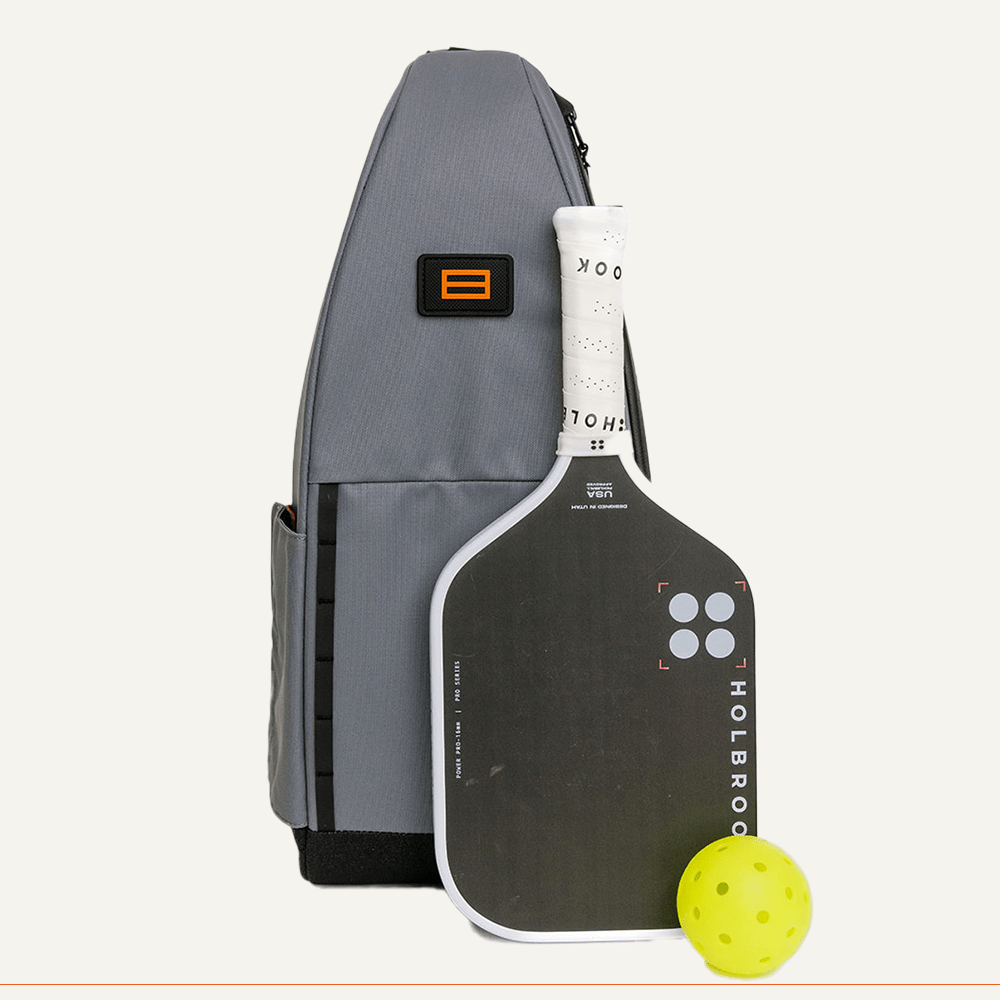 The FORWRD Cross Court Pickleball Sling, a lightweight and durable gray backpack, stands upright—ideal for pickleball. Beside it: a black and white Holbrook paddle with white grip tape and a yellow perforated ball. Set against a plain white background.