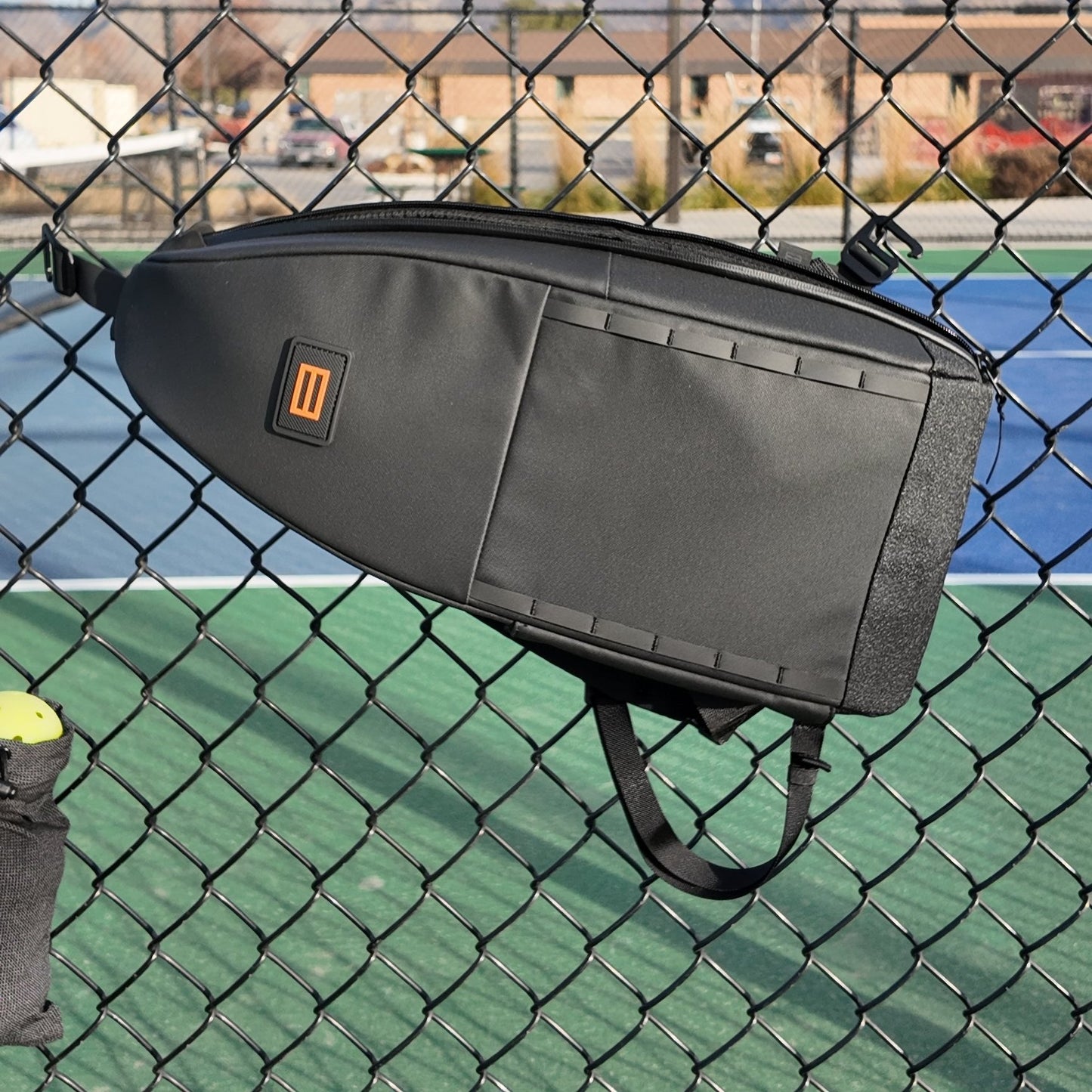 The FORWRD Cross Court Pickleball Sling, a sleek black bag with a rectangular orange and black logo, hangs on a chain-link fence by outdoor courts. Sunlight creates shadows and buildings and trees are visible in the background.