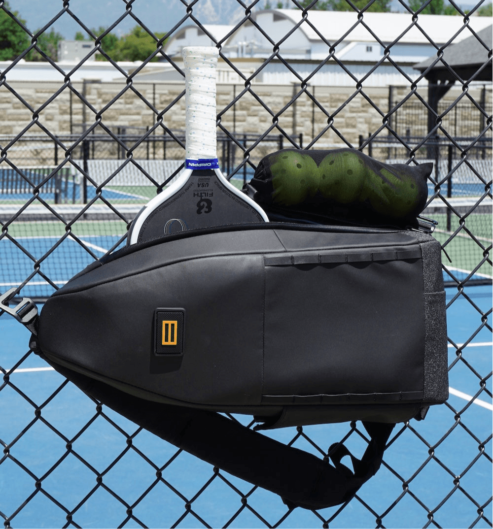 The FORWRD Cross Court Pickleball Sling, featuring a logo with two vertical yellow lines, hangs on a chain-link fence by a blue court with a paddle, towel, and ball peeking out from its lightweight, durable design.