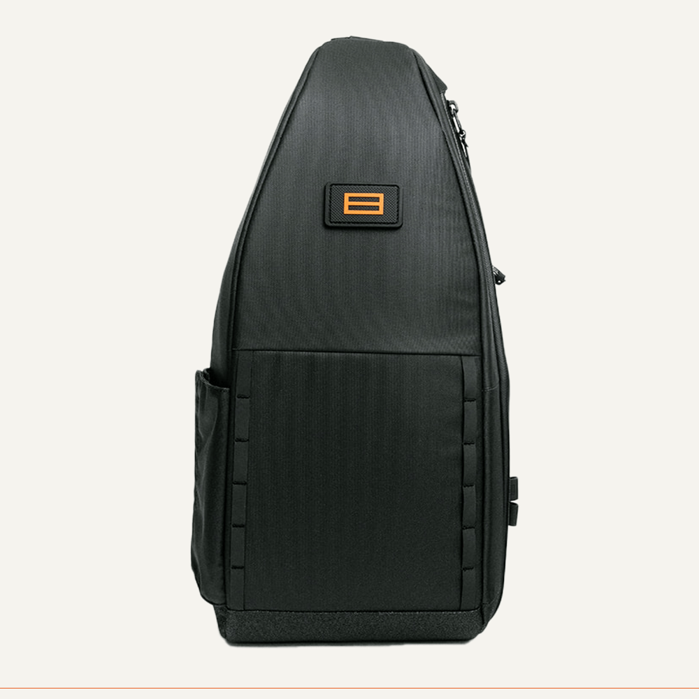 The FORWRD Cross Court Pickleball Sling is a sleek black backpack with an angular, asymmetrical design, side pocket, orange logo near the top, minimalist panel detailing, and stands upright against an off-white background.