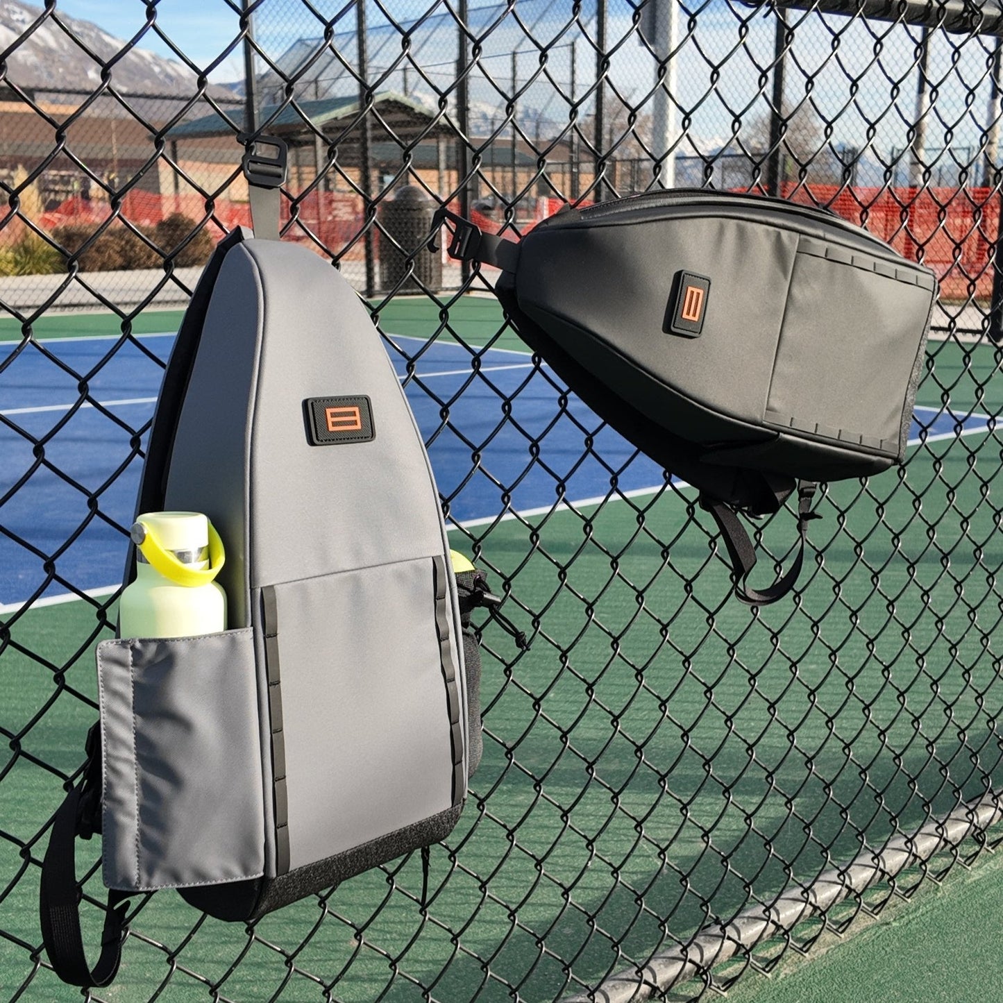 Two gray FORWRD Cross Court Pickleball Sling bags hang on a chain-link fence at a tennis court. One bag, ideal for pickleball gear, holds a yellow water bottle. Blue and green court surfaces meet mountains under partly cloudy skies.