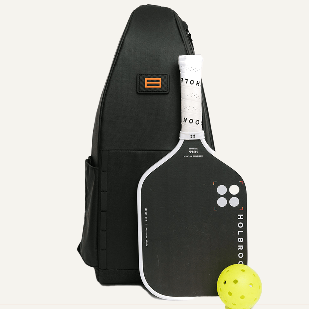 A black FORWRD Cross Court Pickleball Sling stands upright with a lightweight, durable Holbrook paddle—white edge and grip—and a yellow pickleball in front. The bag has side pockets, an orange logo patch, and a light background.