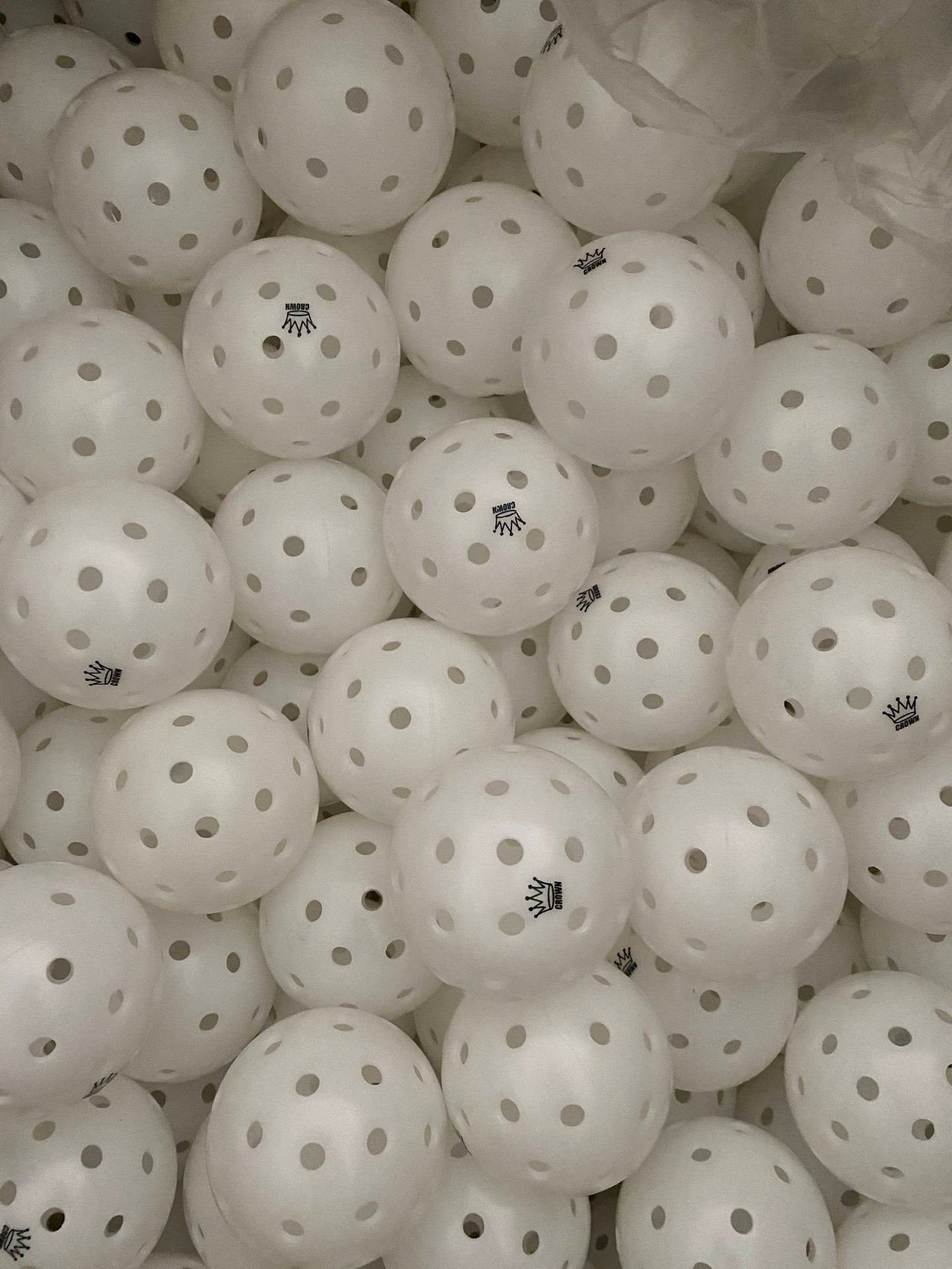 A close-up of Crown Pickleball's High-Performance CONTRAST OUTDOOR Pickleballs (3-PACK COCONUT) shows durable balls with evenly spaced holes, tightly packed and some featuring a black crown logo, filling the frame.