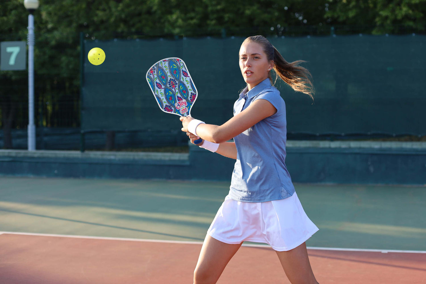 A young woman in a blue shirt and white skirt plays pickleball outdoors, preparing to hit a yellow ball with the Damask 4Joy Pickleball Paddle Premio Series by 4Joy Paddles. Trees and a green fence line the red and green court behind her.