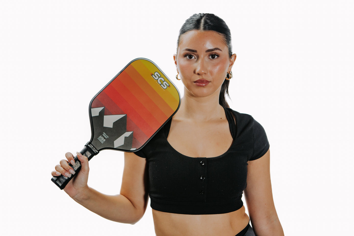 A woman with long dark hair in a ponytail stands against a white background, wearing a black crop top and hoop earrings. She holds the Salted City Sports Darkmode | Evo-Lite Series pro pickleball paddle upright in her right hand.