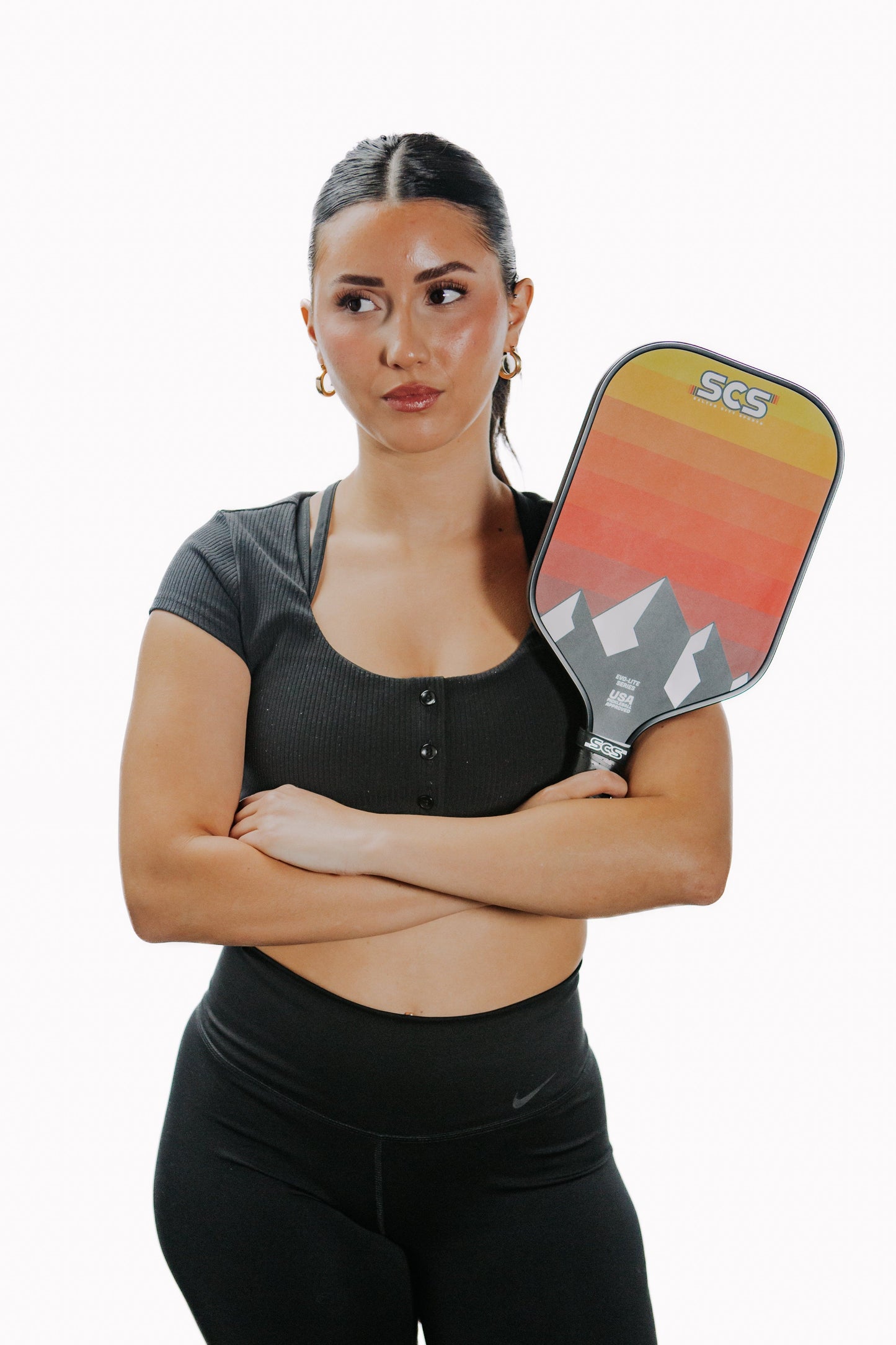A woman with long dark hair and gold hoop earrings wears a black crop top and holds the Salted City Sports Darkmode | Evo-Lite Series pickleball paddle, standing with arms crossed against a plain white background.