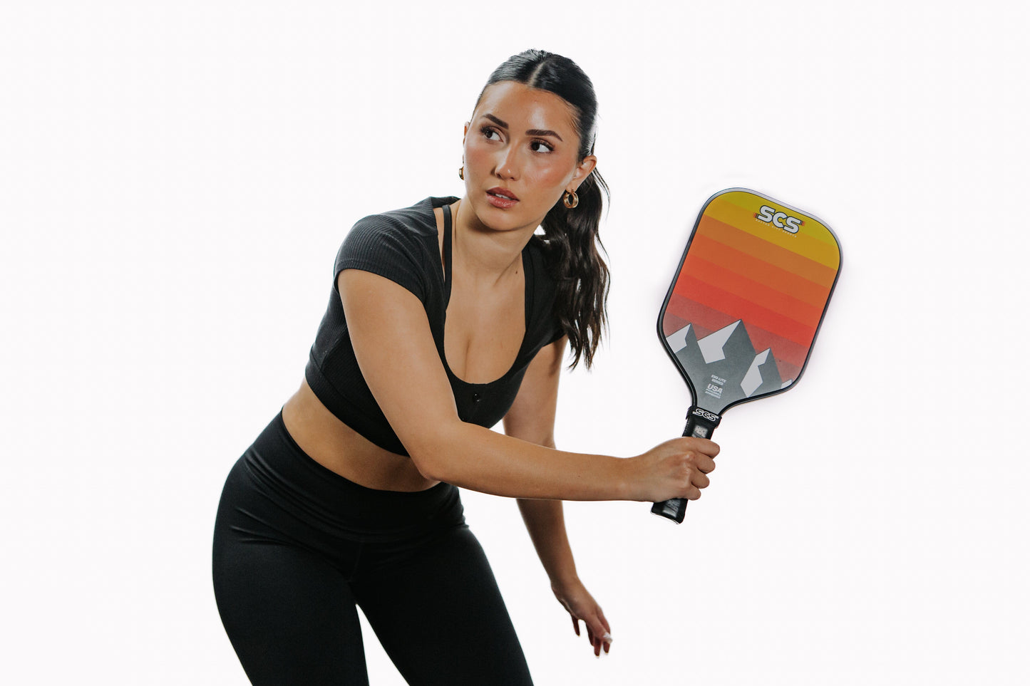 A woman with dark hair in a ponytail, wearing a black crop top and leggings, poses in an athletic stance against a white background holding the Salted City Sports Darkmode | Evo-Lite Series pro-level pickleball paddle with mountain design.